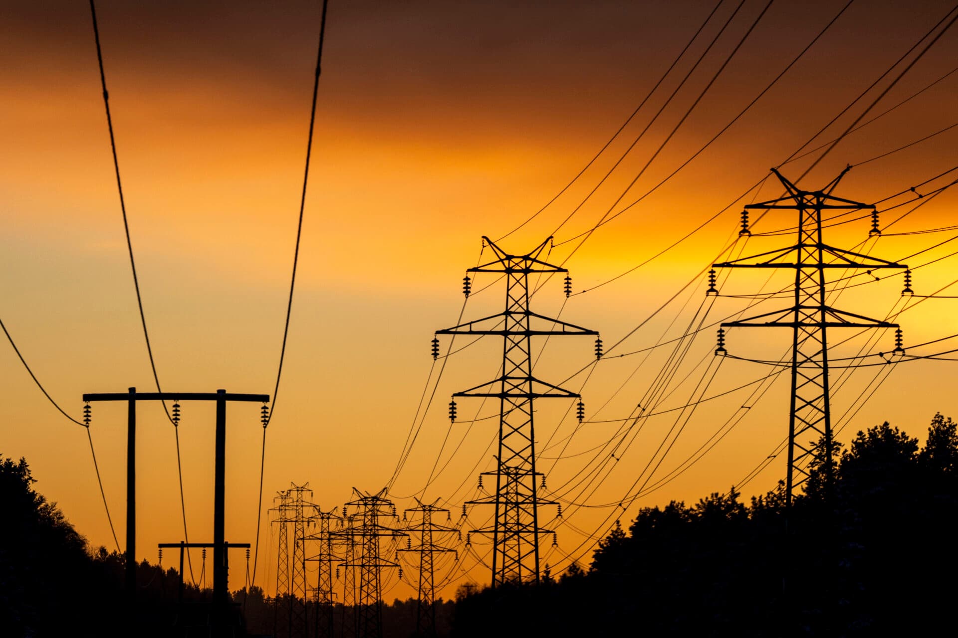 Høyspentledninger og kraftmaster avtegner seg i silhuett mot en oransje og gul solnedgangshimmel, med trær ved foten av mastene.