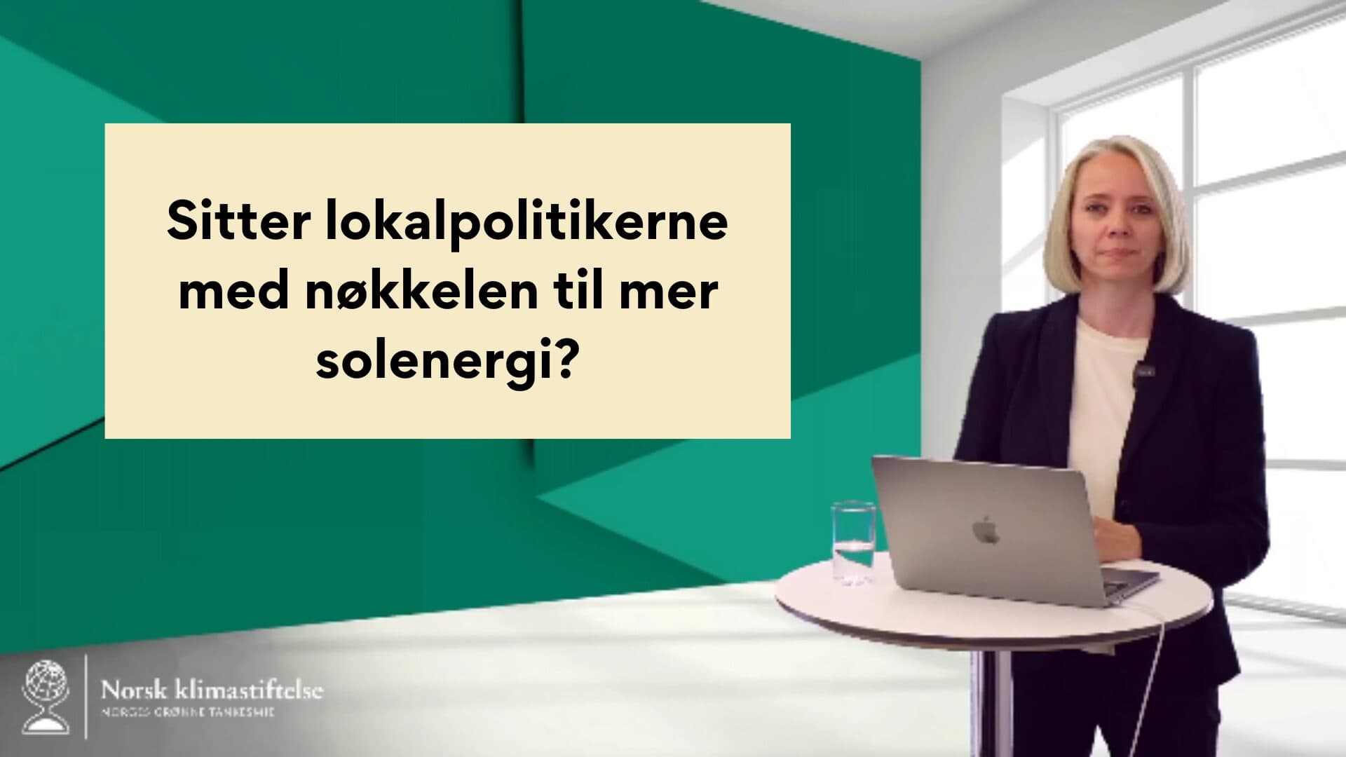 En person står ved siden av et bord med en bærbar datamaskin og et glass vann, sammen med et skilt som leser «Sitter lokalpolitikerne med nøkkelen til mer solenergi?.