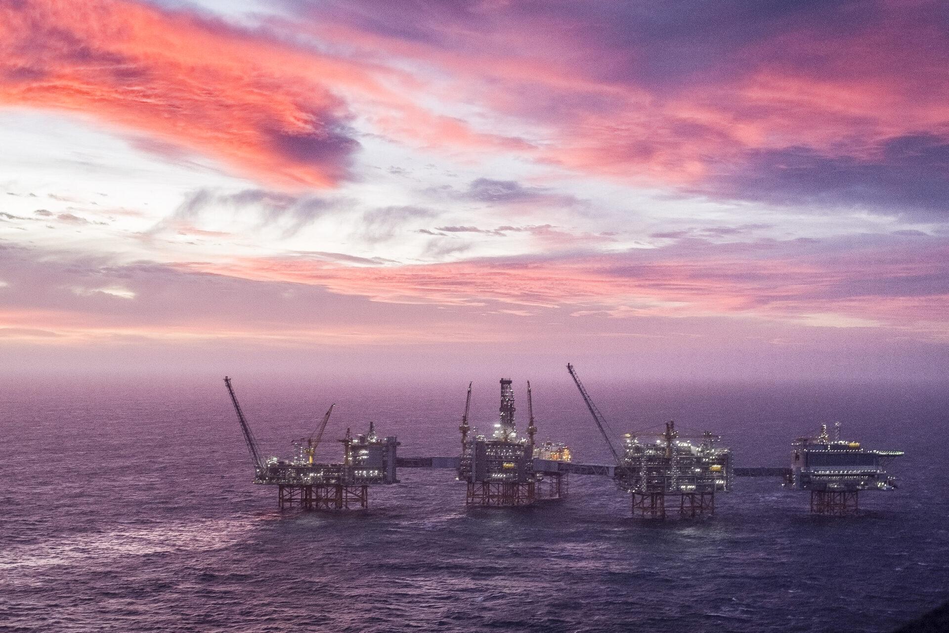 Offshore oljeplattformer på rad på havet under en dramatisk solnedgangshimmel med pulserende skyer.