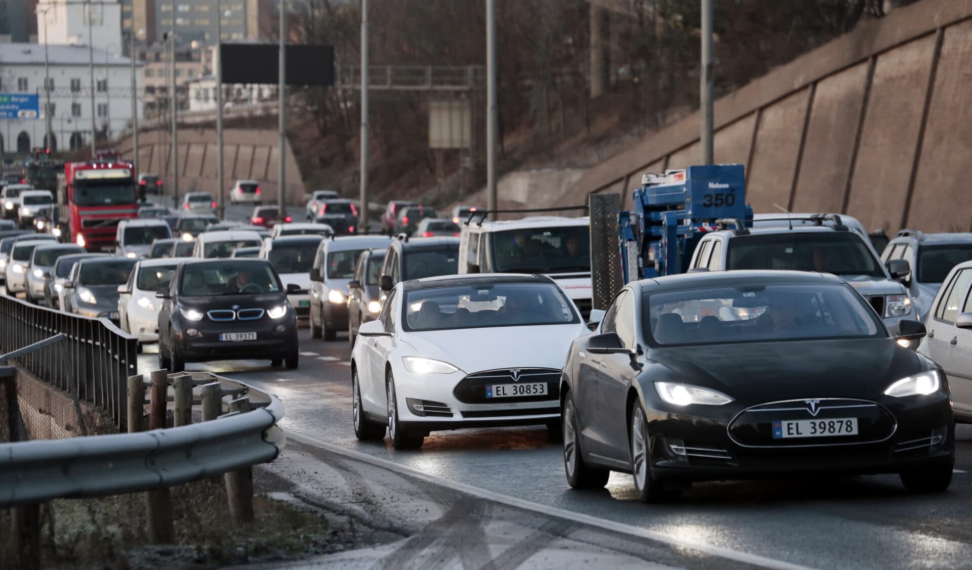 Trafikk på en travel motorvei med flere biler, inkludert Teslaer, i begge retninger. Rekkverk er synlige, og et bybilde er i bakgrunnen.