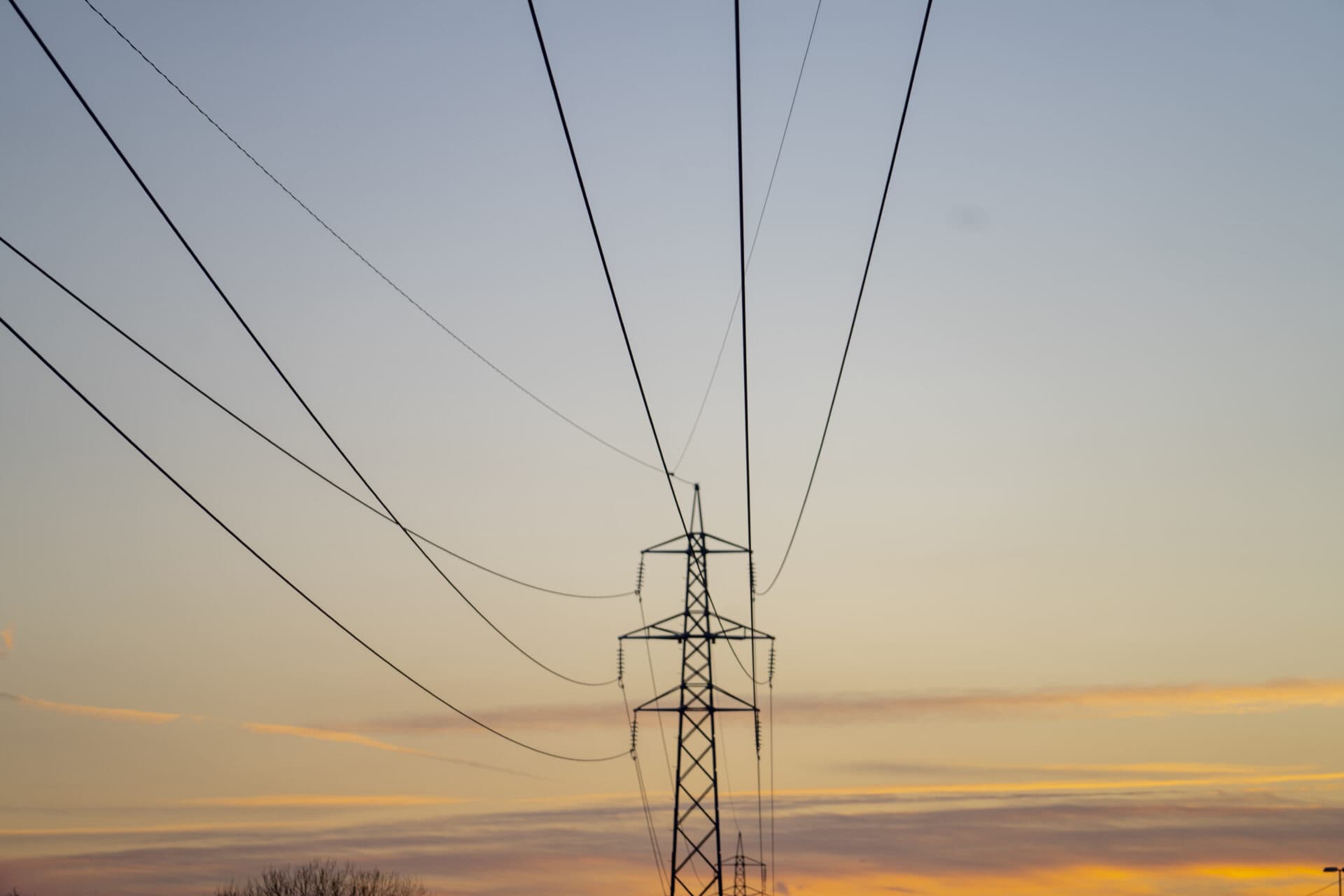 Elektrisk overføringstårn med flere kraftledninger mot en gradienthimmel ved solnedgang.