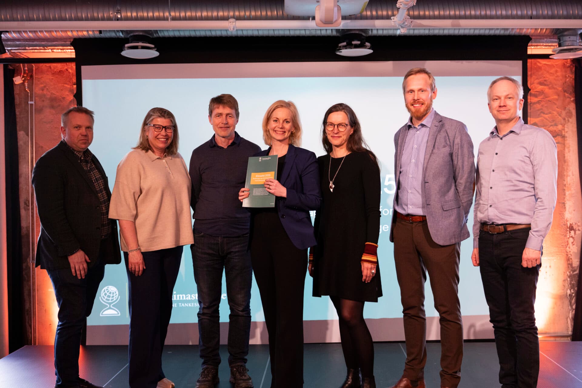 En gruppe på syv personer som står på en scene, med en person som holder en bok og smiler til kameraet.