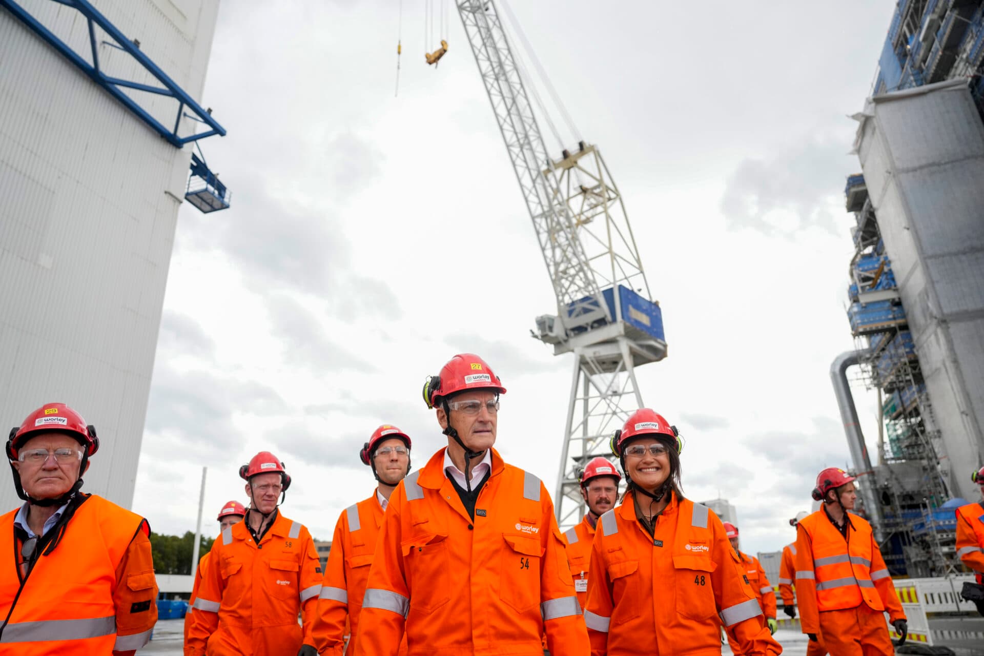 En gruppe mennesker i oransje vernetrøyer og røde hjelmer går rundt på et industriområde med en stor kran i bakgrunnen.