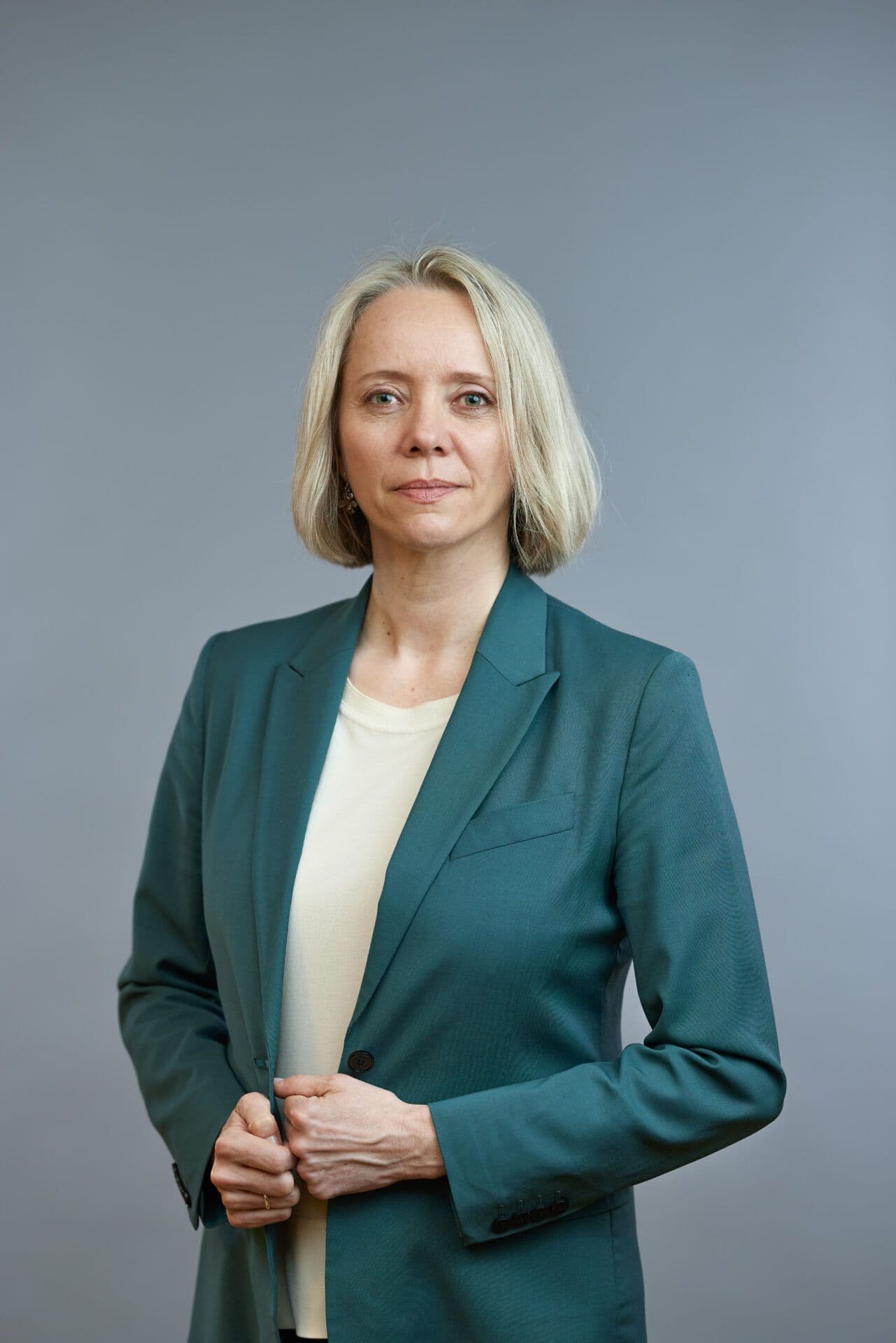 Profesjonell kvinne med blondt hår i en blågrønn blazer over en beige topp, stående mot en grå bakgrunn.