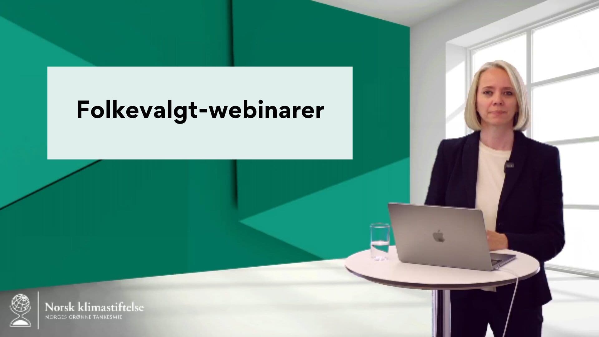 En person står på et podium med en bærbar datamaskin og et glass vann foran. Bakgrunnen har et skilt som sier "Folkevalgt-webinarer." Innstillingen ser ut til å være for en webinarpresentasjon.